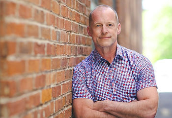 Dave Veale leans against a brick wall, arms crossed, looking directly at the camera.