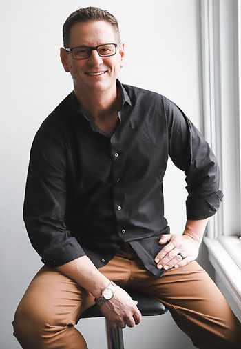 A man in an open dress shirt and casual slacks sits on a stool, looking relaxed, and smiles at the camera.