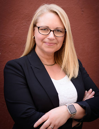A professional woman with blonde hair and glasses smiles slightly, arms across her chest.